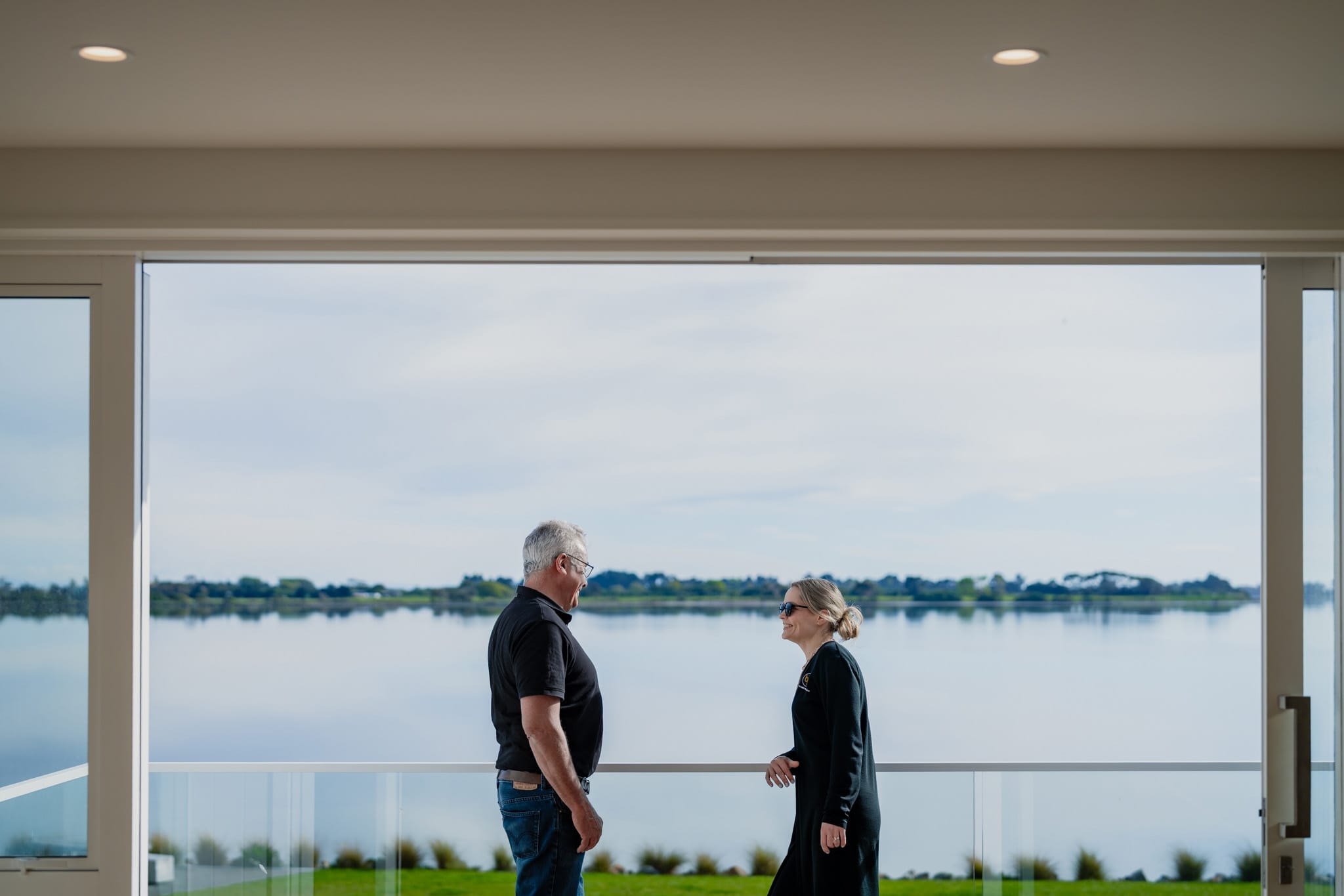 Home builder and client standing on a covered deck overlooking water, discussing a bespoke new build home as part of a design and build residential construction project in New Zealand, showcasing architecturally designed homes, construction management, and new housing developments.