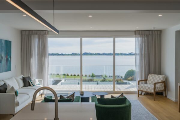 Modern living space with large glazing overlooking water, showcasing architecturally designed homes delivered through design and build and professional home construction in New Zealand.